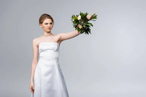 Descontenta novia en vestido blanco mirando flores de boda aisladas en gris - foto de stock