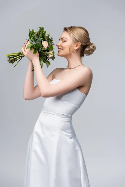 Young and cheerful bride in white dress smelling bouquet of flowers isolated on grey — Stock Photo