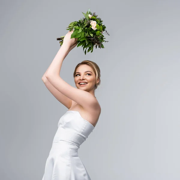 Young and cheerful woman in white wedding dress holding bouquet of flowers above head isolated on grey — Stock Photo