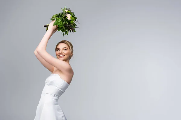Joven y alegre novia en vestido de novia blanco con ramo de flores por encima de la cabeza aislado en gris - foto de stock
