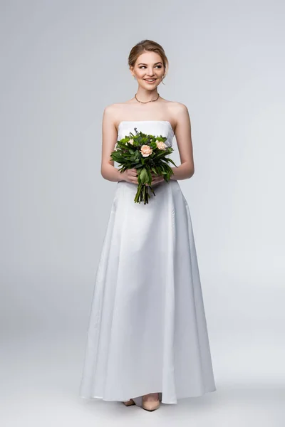 Sposa sorridente in abito da sposa bianco con mazzo di fiori su grigio — Foto stock