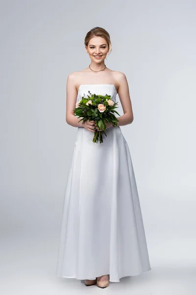 Fille souriante en robe de mariée blanche tenant bouquet de fleurs sur gris — Photo de stock