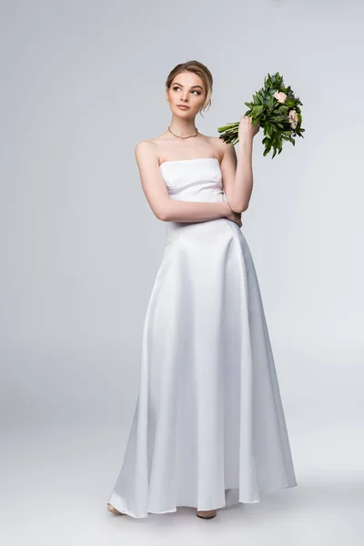 Pensive girl in white wedding dress holding bouquet of flowers on grey — Stock Photo