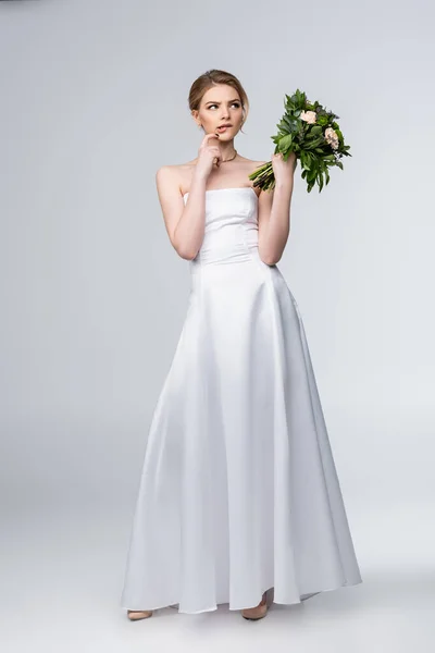 Pensive girl in white wedding dress holding bouquet of flowers and touching face on grey — Stock Photo