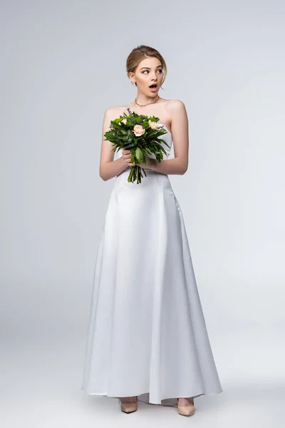 Shocked girl in white wedding dress holding bouquet of flowers isolated on grey — Stock Photo