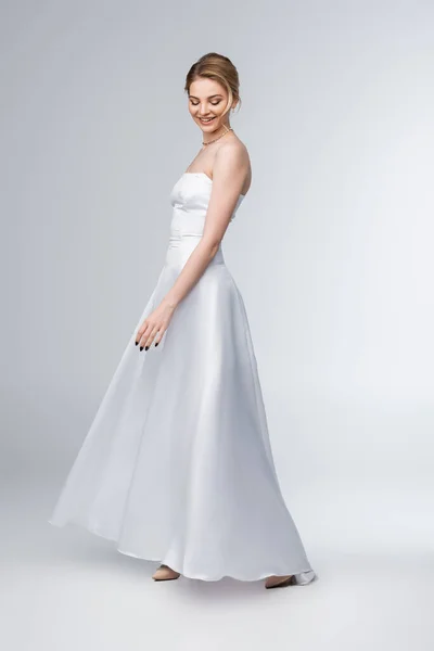 Улыбающаяся невеста в элегантном свадебном платье, стоящем на сером — стоковое фото