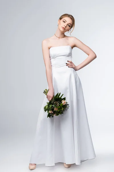 Bride in elegant wedding dress holding bouquet of flowers and standing with hand on hip on grey — Stock Photo