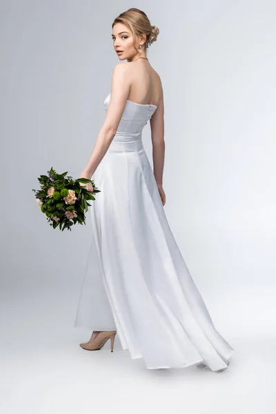 Mariée attrayante dans une élégante robe de mariée tenant bouquet de fleurs sur gris — Photo de stock