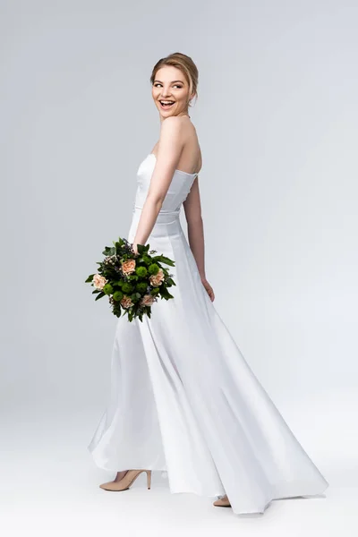 Attractive bride in elegant wedding dress holding bouquet of flowers and smiling on grey — Stock Photo