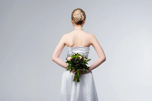 Vista trasera de la novia en vestido de novia elegante celebración ramo de flores detrás de la espalda aislado en gris - foto de stock