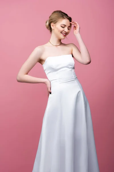 Happy young bride in white wedding dress standing with hand on hip isolated on pink — Stock Photo