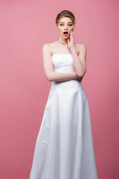 Novia sorprendida en vestido de novia blanco tocando la cara aislada en rosa - foto de stock