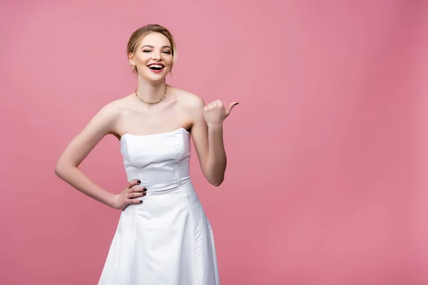 Sposa felice in abito da sposa bianco puntando con il pollice e ridendo sul rosa — Foto stock