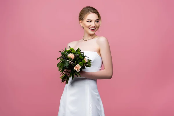 Novia sonriente en vestido de novia blanco sosteniendo flores aisladas en rosa - foto de stock