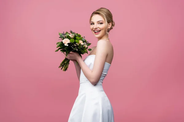 Cheerful bride in white wedding dress holding flowers isolated on pink — Stock Photo