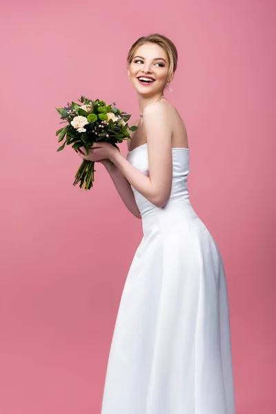 Mariée gaie en robe de mariée blanche tenant des fleurs et regardant loin isolé sur rose — Photo de stock