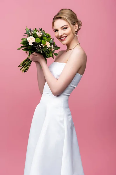 Novia alegre en vestido de novia blanco sosteniendo flores y mirando a la cámara aislada en rosa - foto de stock