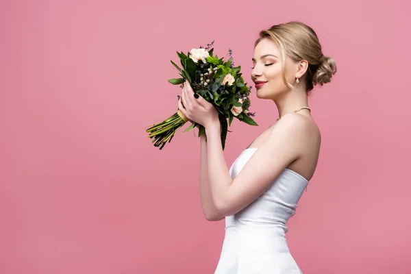 Happy bride in white wedding dress smelling flowers isolated on pink — Stock Photo