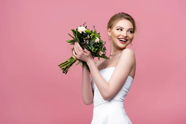 Alegre chica en vestido de novia blanco sosteniendo flores y mirando hacia otro lado aislado en rosa - foto de stock