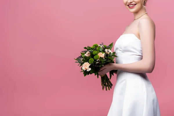 Abgeschnittene Ansicht einer glücklichen Braut im weißen Hochzeitskleid mit Blumen auf rosa — Stockfoto