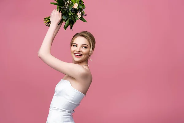 Happy bride in white wedding dress holding flowers above head isolated on pink — Stock Photo