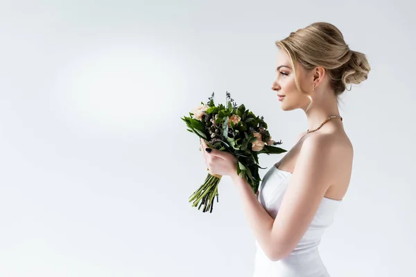 Vista laterale della sposa attraente che tiene fiori di nozze su bianco — Foto stock