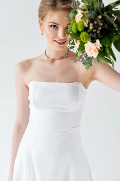Novia joven y feliz en vestido de novia elegante con flores aisladas en blanco - foto de stock