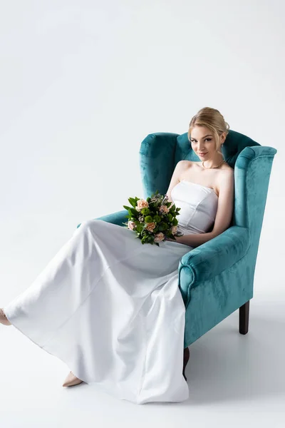 Attractive bride in elegant wedding dress holding flowers while sitting in armchair on white — Stock Photo