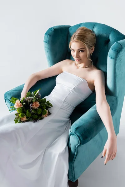 Belle mariée dans une élégante robe de mariée tenant des fleurs tout en étant assis dans un fauteuil sur blanc — Photo de stock