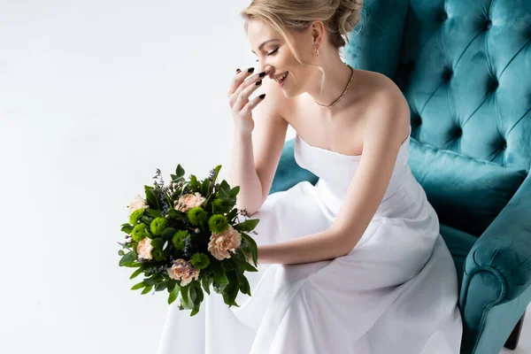 Mariée souriante dans une élégante robe de mariée tenant des fleurs tout en étant assis dans un fauteuil et toucher le visage isolé sur blanc — Photo de stock