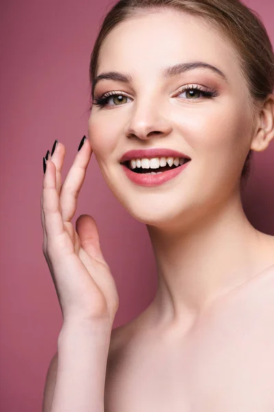 Mujer joven desnuda con sombra de ojos y lápiz labial tocando la cara y sonriendo en rosa - foto de stock