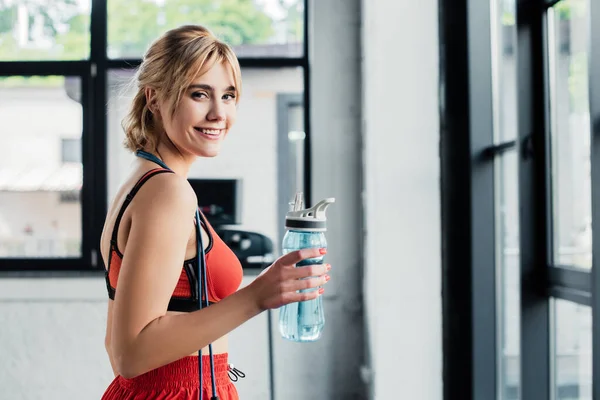 Cheerful sportswoman holding sports bottle with water in gym — Stock Photo