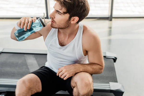 Tired sportsman drinking water and holding sports bottle while sitting on treadmill — Stock Photo