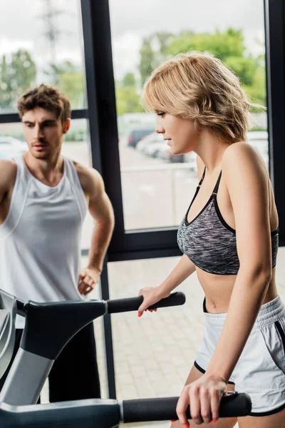 Selective focus of attractive sportswoman touching handrails on treadmill near handsome sportsman — Stock Photo