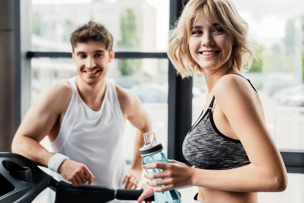 Selective focus of happy sportswoman holding sports bottle with water near man in gym — Stock Photo