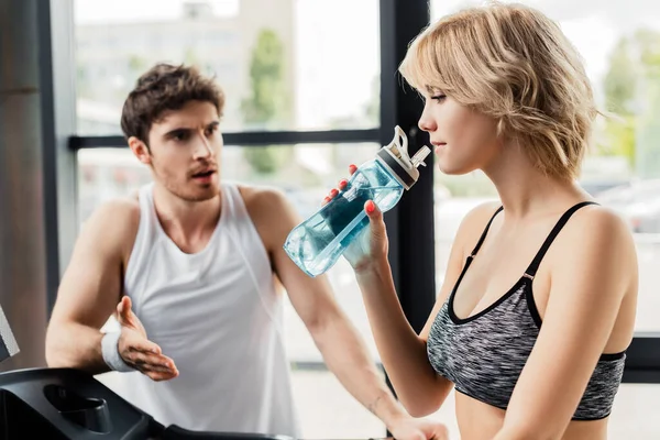 Foyer sélectif d'attrayant sportif tenant bouteille de sport avec de l'eau près de l'homme dans la salle de gym — Photo de stock