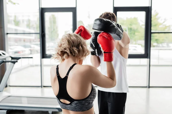 Vista posterior de la pareja deportiva en guantes de boxeo y almohadillas de ejercicio en el gimnasio - foto de stock