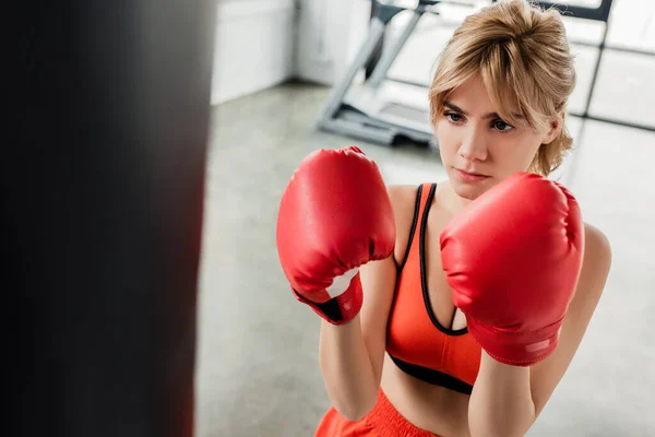 Selective focus of young sportswoman in boxing gloves looking at punching bag — Stock Photo