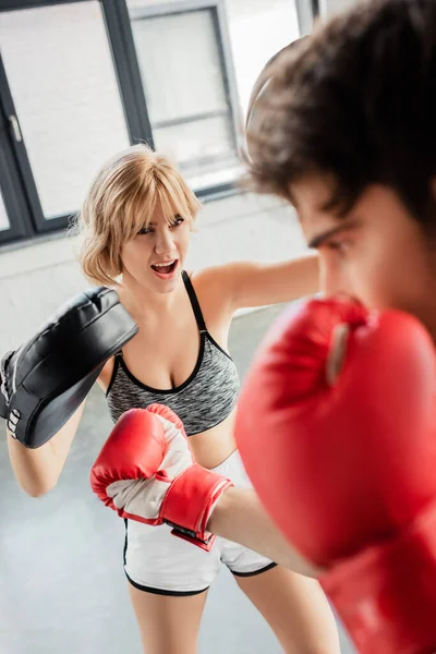 Enfoque selectivo de chica emocional en almohadillas de boxeo ejercicio con deportista en el gimnasio - foto de stock