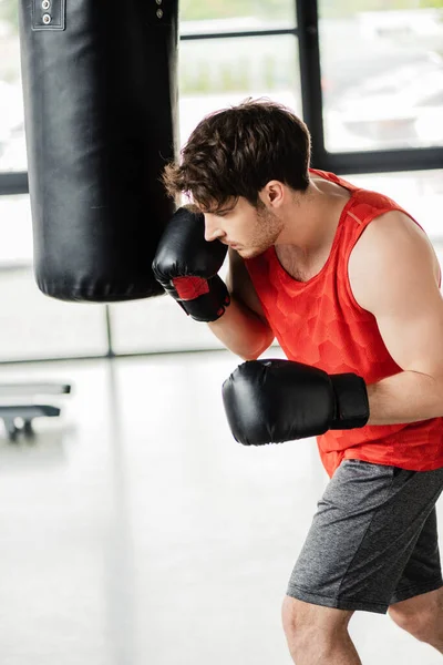 Uomo atletico in abbigliamento sportivo e guanti da boxe che si allena con il sacco da boxe — Foto stock