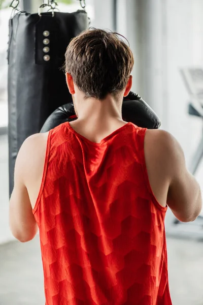 Vista trasera del hombre atlético en ropa deportiva y guantes de boxeo trabajando con el saco de boxeo - foto de stock