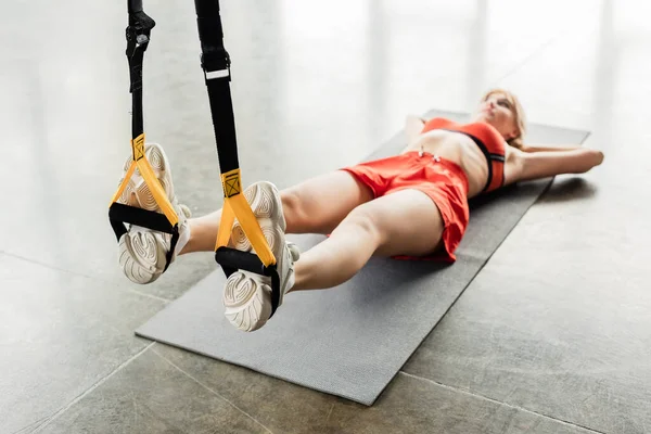 Foco seletivo da jovem atlética em roupas esportivas exercitando-se com bandas de resistência enquanto deitado no tapete de fitness — Fotografia de Stock