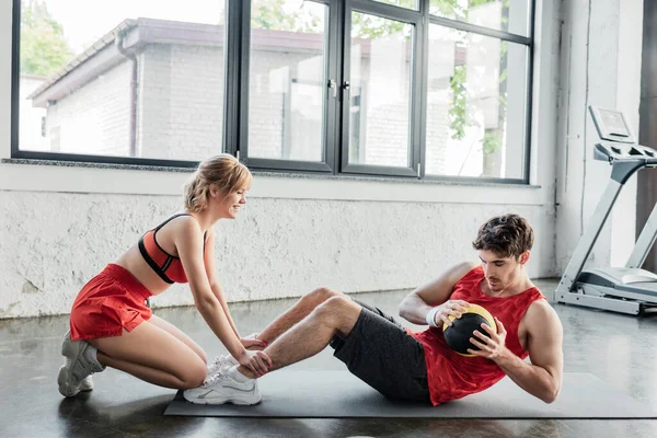 Glückliches Mädchen, das Beine eines Sportlers hält, der mit Ball auf Fitnessmatte trainiert — Stockfoto