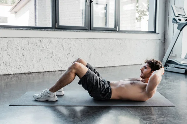Vista lateral del deportista sin camisa haciendo sentadas en la colchoneta de fitness en el gimnasio - foto de stock