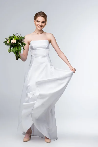 Cheerful bride touching elegant wedding dress and holding flowers on white — Stock Photo
