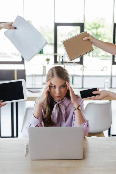 Femme d'affaires stressée assise près de collègues tenant des gadgets avec écran blanc, ordinateur portable et papiers — Photo de stock