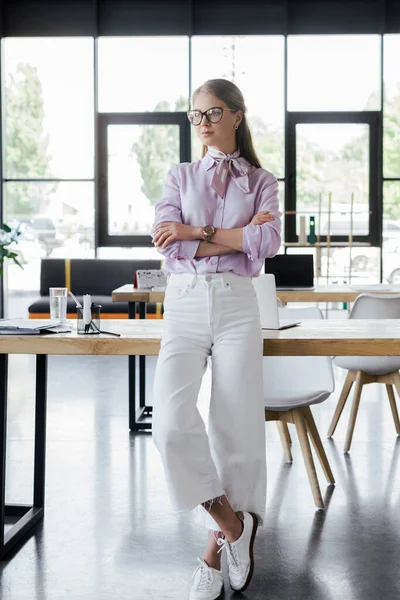 Hermosa mujer de negocios en gafas de pie con los brazos cruzados en la oficina - foto de stock