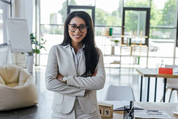 Happy businesswoman in glasses standing with crossed arms near table in office — Stock Photo