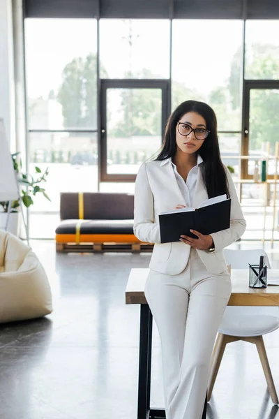 Attrayant femme d'affaires tenant un cahier et regardant la caméra dans le bureau — Photo de stock