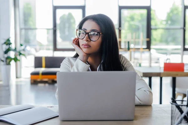 Sad businesswoman in glasses looking away near laptop and notebook on table — Stock Photo